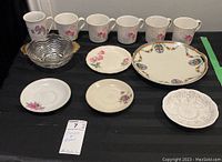 Twelve pieces of assorted china and glass including six mugs, three saucers, one side plate, one Noritake platter, and one glass bowl with gold trim, displayed on black background.