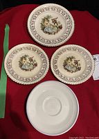 Four assorted plates and saucers arranged on red cloth, showing ornate floral border and pastoral scene design.