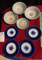 Eight assorted pieces of china on red cloth, showing four pale green rimmed plates with floral centers, three blue rimmed bowls, one matching saucer.