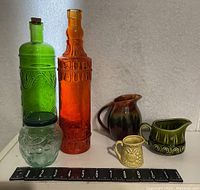 Group view of six decorative glass and pottery items showing bottles, jar, and jugs with sizing scale