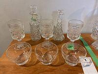 Group shot of two pressed glass cruets, three matching wine glasses, and three fruit nappies arranged on a wooden surface.