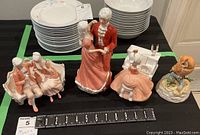 Four figurines displayed on black surface with ruler showing size: couple on loveseat Peach Elegance, woman at piano Fascination, dancing couple, and owl figurine