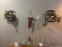 Two black metal wall-mounted plant holders with white ceramic pots containing green ivy faux plants, and the bamboo wind chime hanging between them
