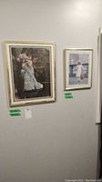 Two framed prints hung on a wall, one larger with a girl in blue dress holding flowers, one smaller with a girl in white dress and hat.