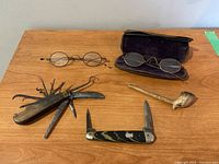 Photo showing two pairs of wire-framed spectacles (one inside a lined metal case), two pocket knives with multiple tools, and a bone or antler pipe with chip