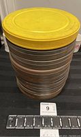 Stack of nine metal film canisters with a yellow plastic lid on top, showing size reference with a measuring tape.