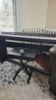 Casio Privia digital keyboard setup next to a radiator, showing full keyboard and folding stool below it. A cable and foot pedal are also visible on the padded seat of the stool.