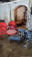 Photo showing two red metal chairs, small round red metal table, blue stools and footstool in basement environment.