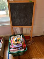Wooden easel with chalkboard side showing chalk markings and a basket placed underneath filled with assorted children's books.