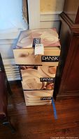 Three stacked original boxes of Dansk Kitchen Santiago white dinnerware, unopened and stacked on the floor near wooden furniture.