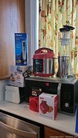 Full lot overview showing all kitchen appliances including the boxed electric wine opener, pressure cooker, blender, toaster oven, mini waffle maker box, sandwich maker box, and foil cutter box on countertop under window with floral curtains.