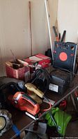 View of multiple tools on a table including Black & Decker cordless hedge saw, yellow finishing sander, air compressor, hardware boxes, and ladder shelf in background