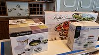 Photo showing three boxed sets on countertop including Fagor cookware set, microwave cookware set, and boxed 3-tier buffet server.