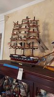 Full view of the wooden ship model showing multiple sails, painted hull, and black stand base.