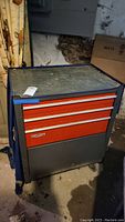 Full front view of Craftsman tool chest on wheels in basement, showing red and gray metal body with aluminum handles and visible dirt on top surface.
