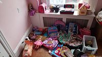 Room corner with multiple items including backpacks, dolls, toy cash register, dollhouse, and purses spread on floor and window ledge