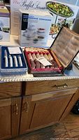 View of the 3-tier buffet server box, microwave cookware set box, stag horn handle utensil set in case with foil packaging, and a large hammered aluminum tray on top of a wooden cabinet.