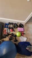 Shows various dumbbells, kettlebell, exercise balls, roller wheel and resistance bands placed on floor with wall in background.