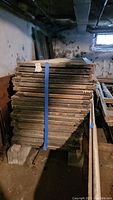 Stack of vintage wooden windows bound with blue strap, showing aged wood and paint chipping, stored in basement.