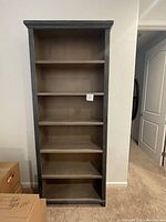 Frontal view of the gray-blue wooden bookcase showing all six fixed shelves and top decorative molding, placed against beige wall.