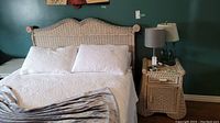 Wicker queen headboard and bed frame fully assembled with white bedding, matching bedside table with lamp and books.