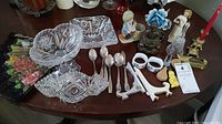 Wide shot of table showing lot contents including brass candle holders with candles, glass bell, crystal candy dish, glass ashtray, serving utensils, napkin holders and Willowtree figurine