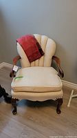 Front view of vintage upholstered armchair showing cream fabric upholstery, carved wooden arms and legs, with a red throw on the backrest.