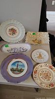 Six various plates and dishes on a table including floral, scenic, and illustration designs.