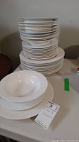 Stack of white dinner plates, dessert plates and bowls arranged on a table with a removal tag visible