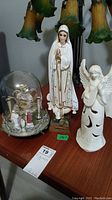 Photo showing a 14 inch religious figurine with a medallion, a ceramic angel figurine, and miniature figurines under a glass dome.