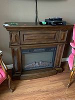 Front view of the electric fireplace showing dark wood mantel with panel detailing, glass front fireplace insert, simulated logs, and remote on top.