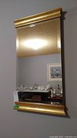 Full view of a large bevelled glass mirror mounted on a wall, gold frame top and bottom visible, reflection of ceiling light and a room with furniture and dishes.