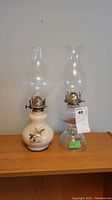 Photo showing two vintage oil lamps on wooden surface, one ceramic with bird design and one clear glass base.