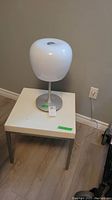 Side angle showing white square table with metal legs and rounded white lamp on top against beige wall