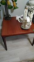 Full view of the wooden square table with a polished brown finish and tapered legs, placed on a light-colored floor with some items on top removed for lot sale.