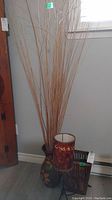 Photo showing pottery vase with tall natural reeds, adjacent lamp and rattan basket on floor near wall.