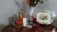 Items include a white floral vase, Mason's decorative plate, copper pitcher, coffee carafe, multiple candles, and glass decorative items displayed on a wooden surface
