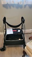 Nexus B Walker rollator from front showing padded seat, backrest, wheels and storage bag, with manual on seat.