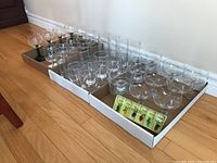 Boxes containing assorted glass stemware set including wine glasses with green stems, clear highball glasses, dessert cups, and wine charms.