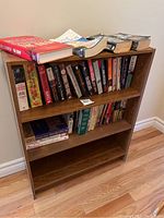 Wooden bookshelf with novels on three shelves. Books include paperback and hardcover editions, genres mostly fiction.