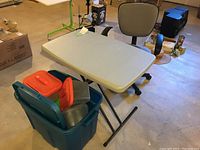 Photo showing small folding table with beige top and black legs next to teal plastic storage tote filled with assorted smaller totes with various colored lids.