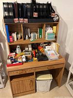 Full view of oak desk with shelving filled with black plastic magazine holders, assorted office supplies on desktop including two telephones, calculators, flashlights, office tools, and a paper shredder below the desk.