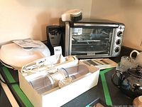 Full view of toaster oven, George Foreman grill, can opener, immersion blender, chopper/slicer, and grater arranged on countertop.