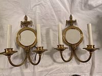 Pair of vintage brass wall sconces with two candle fixtures each and central round mirrors, displayed on fabric background.