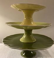 Three stacked ceramic cake plates in yellow, light green, and dark green, showing decorative scalloped edges and pedestal bases.