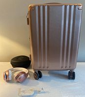 Calpak rose gold carry-on suitcase with over-ear headphones, carrying case and luggage tag arranged side by side.