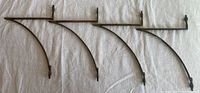 Four curved metal brackets with two screw holes on each, laid side-by-side on a white cloth background. Brackets have a dark antique brass finish and are visibly new and unused.