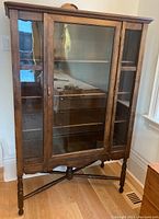 Front view of an antique wooden display cabinet with central glass door and glass side panels, three interior shelves visible. Detailed woodwork on top and legs.