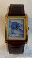 Front view of watch showing the 5 cent Canada Post wildlife stamp face with bear design in blue on rectangular gold-tone case and brown leather strap.