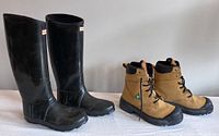 Pair of black Hunter insulated rain boots and tan Aggressor steel toe boots side by side on table.